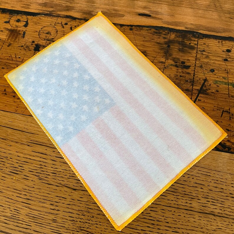 Pu&ograve; includere: Un toppa di bandiera americana in tessuto bianco con un bordo giallo. La bandiera &egrave; sbiadita e ha un aspetto vintage.