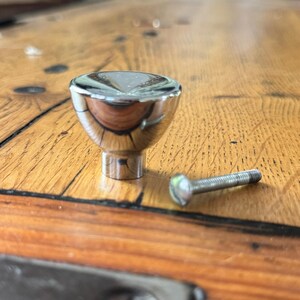 Puede incluir: Un pomo para mueble de cromo pulido con una parte superior lisa y redondeada y una base cilíndrica, acompañado de un tornillo de montaje metálico a juego. El herraje presenta un acabado altamente reflectante y similar a un espejo.