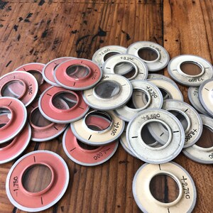 以下が含まれることがあります： 木製の表面に、丸い白とピンクの金属製の円形のレンズのような物体が数字とプラスまたはマイナスの記号で積み重ねられています。