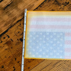 Pu&ograve; includere: Un primo piano di una bandiera americana in tessuto bianco con cuciture gialle attorno ai bordi. La bandiera viene misurata con un metro a nastro, mostrando che &egrave; larga circa 15 cm.