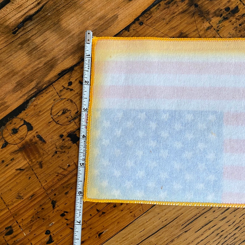 Pu&ograve; includere: Un primo piano di una bandiera americana in tessuto bianco con cuciture gialle attorno ai bordi. La bandiera viene misurata con un metro a nastro, mostrando che &egrave; larga circa 15 cm.
