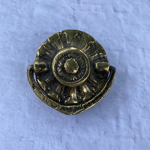 Puede incluir: Un pomo de armario de metal dorado con un diseño decorativo y ornamentado. El pomo tiene una forma circular con un centro elevado y una manija curva abierta.