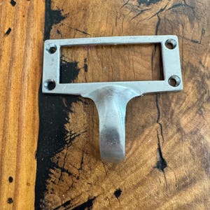 May include: A silver-colored metal label holder with a rectangular frame and a curved handle. The frame has four holes for mounting. The label holder is designed to hold a small label or tag.