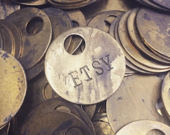 May include: A close-up of a pile of brass-colored metal tags with a single tag in the center that reads "ETSY".
