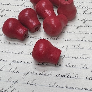 May include: A group of seven red wooden knobs with a rounded top and a small hole in the center. The knobs are arranged in a random pattern on a white surface with handwritten text.