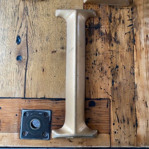 May include: A close-up of a large, golden, metal letter "I" mounted on a wooden surface. The letter is made of a thick, textured metal and has a slightly rounded top.