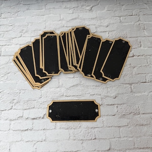 May include: A collection of black rectangular metal nameplates with gold trim, arranged on a white brick surface. Each plate has two holes for mounting. The plates are of varying quantities, with some stacked and others spread out.