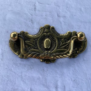 Può includere: Una maniglia per cassetto in ottone vintage con un design ornamentale. La maniglia ha un manico curvo e due pomelli rotondi su ciascuna estremità.