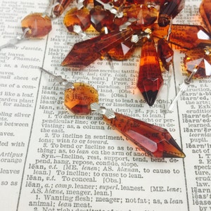 May include: A close-up of a group of amber glass crystal prisms with silver metal tops. The prisms are scattered on a page of a dictionary.
