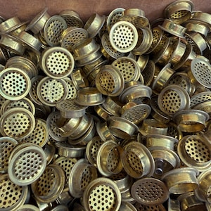 May include: A pile of small, round, brass-colored vents with a perforated surface. The vents have a circular design with a raised outer rim and a central, recessed area filled with small holes. The overall appearance suggests a hardware or industrial component.