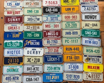 May include: A collection of vintage license plates from different states in the United States. The plates are arranged in a grid pattern on a wooden surface. Each plate features a unique design and state abbreviation. The plates are in various colors, including blue, green, red, yellow, and white.