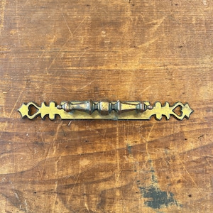 Puede incluir: Un tirador de cajón de metal dorado con un diseño en forma de corazón en cada extremo. El tirador tiene un diseño decorativo y ornamentado.