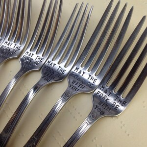 May include: Five silver forks with the phrase "May the fork be with you" engraved on each. The forks are arranged in a row on a light beige surface.