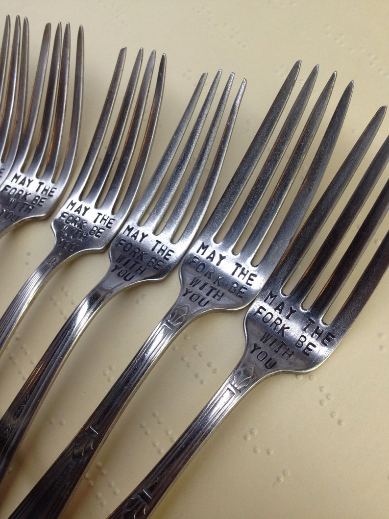 May include: Five silver forks with the phrase "May the fork be with you" engraved on each. The forks are arranged in a row on a light beige surface.