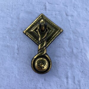May include: A gold-toned metal cabinet pull with a decorative, twisted design. The pull is attached to a square base with a diamond pattern.