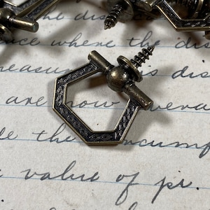 May include: A close-up of a hexagonal-shaped, antique brass, decorative hardware with a screw and a small knob. The hardware has a decorative pattern on the surface.