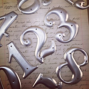 May include: A collection of silver metal house numbers with a decorative, ornate design. The numbers include 1, 2, 3, 4, 8, and a question mark.