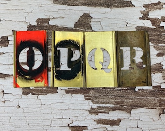 May include: Four metal stencils with the letters O, P, Q, and R cut out. The stencils are arranged in a row, with the O stencil on the left and the R stencil on the right. The stencils are on a white wooden background.