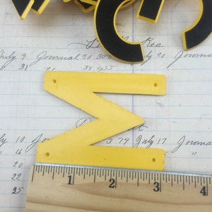 May include: A yellow plastic letter Z with black trim and two holes. The letter is sitting on a piece of paper with handwritten text and a wooden ruler with inch markings.