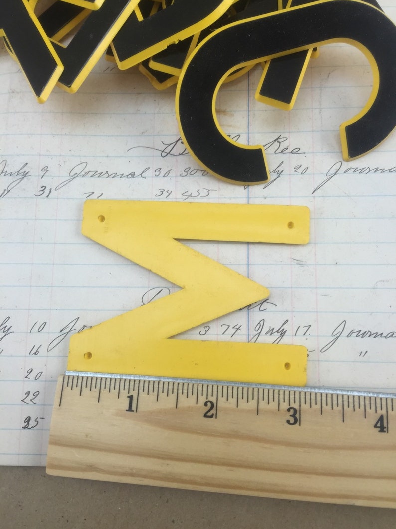 May include: A yellow plastic letter Z with black trim and two holes. The letter is sitting on a piece of paper with handwritten text and a wooden ruler with inch markings.