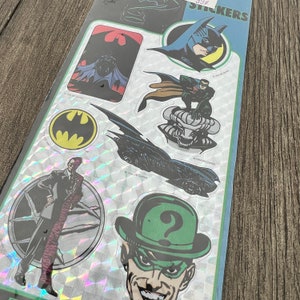Peut inclure: Une feuille d'autocollants holographiques repr&eacute;sentant des personnages de l'univers Batman. Les autocollants comprennent Batman, Robin, l'Homme-Myst&egrave;re, Double-Face et la Batmobile. Les autocollants sont tous en noir et blanc avec une finition holographique.
