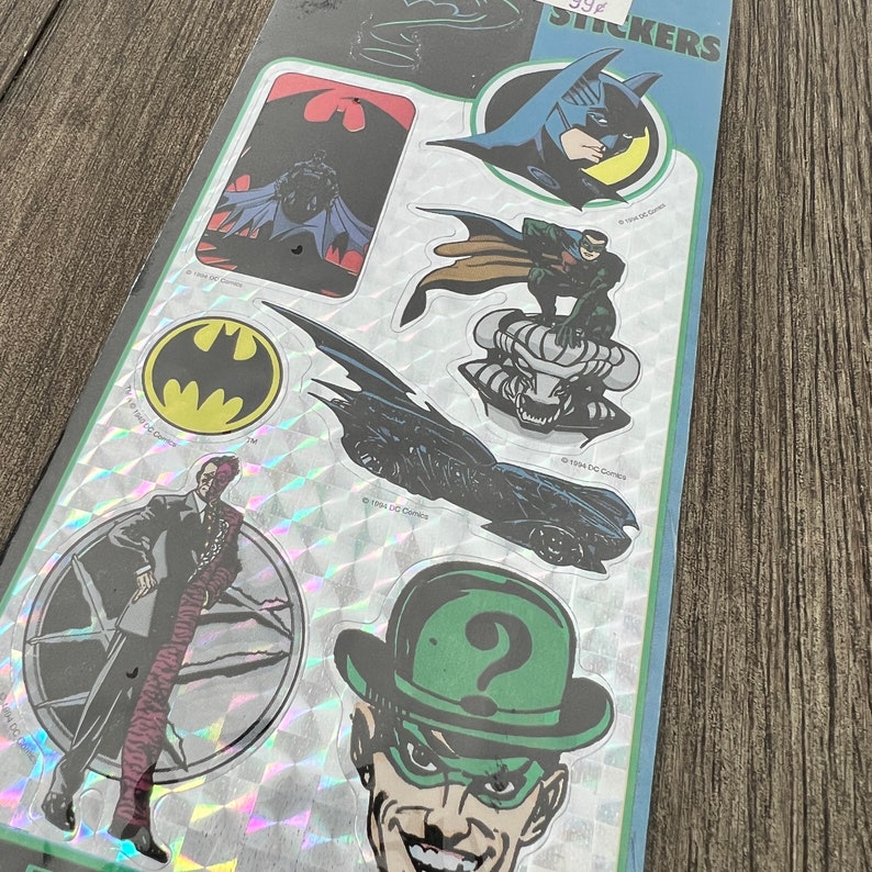 Peut inclure: Une feuille d'autocollants holographiques repr&eacute;sentant des personnages de l'univers Batman. Les autocollants comprennent Batman, Robin, l'Homme-Myst&egrave;re, Double-Face et la Batmobile. Les autocollants sont tous en noir et blanc avec une finition holographique.