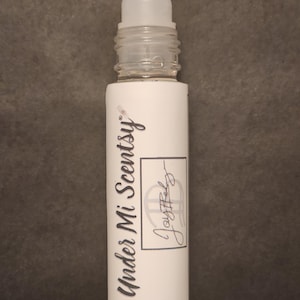 Puede incluir: Un frasco roll-on cil&iacute;ndrico con tapa de pl&aacute;stico transparente y etiqueta blanca. La etiqueta presenta el texto Under Mi Scentsy en una fuente cursiva oscura, junto a un recuadro rectangular que contiene el texto manuscrito Joyful.
