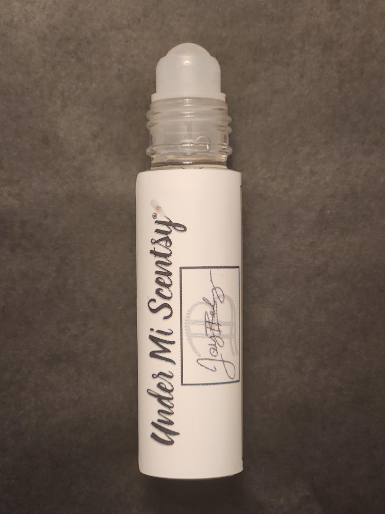 Puede incluir: Un frasco roll-on cil&iacute;ndrico con tapa de pl&aacute;stico transparente y etiqueta blanca. La etiqueta presenta el texto Under Mi Scentsy en una fuente cursiva oscura, junto a un recuadro rectangular que contiene el texto manuscrito Joyful.