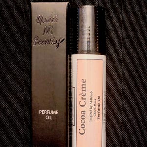 Puede incluir: Una caja rectangular negra de aceite de perfume etiquetada Under Mi Scentsy y 10 ml (0,33 fl oz) junto a una botella cil&iacute;ndrica de aceite de perfume con una etiqueta rosa claro que dice Cocoa Cr&egrave;me, inspirado en Al-Rehab Choco Musk, 10 ml.