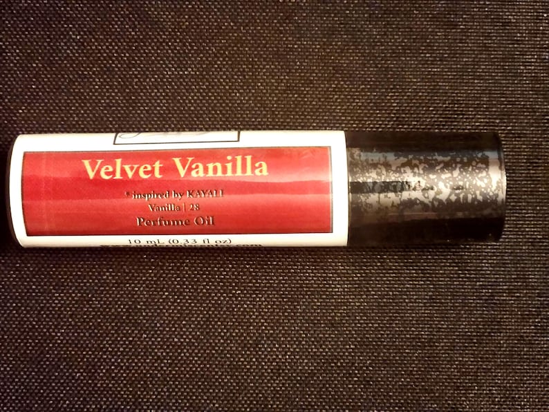 May include: A cylindrical perfume oil roller bottle with a white label featuring a red rectangular accent. The text reads Velvet Vanilla, inspired by KAYALI Vanilla | 28, Perfume Oil, and 10 ml (0.33 fl oz). The cap is black with a textured finish.