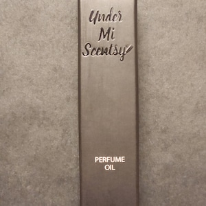 Puede incluir: Una caja rectangular de color marr&oacute;n oscuro para aceite de perfume. El frente presenta el texto en relieve Under Mi Scentsy en una fuente cursiva, seguido del texto impreso PERFUME OIL y la medida de volumen 10mL (0.33 fl oz) en la parte inferior.