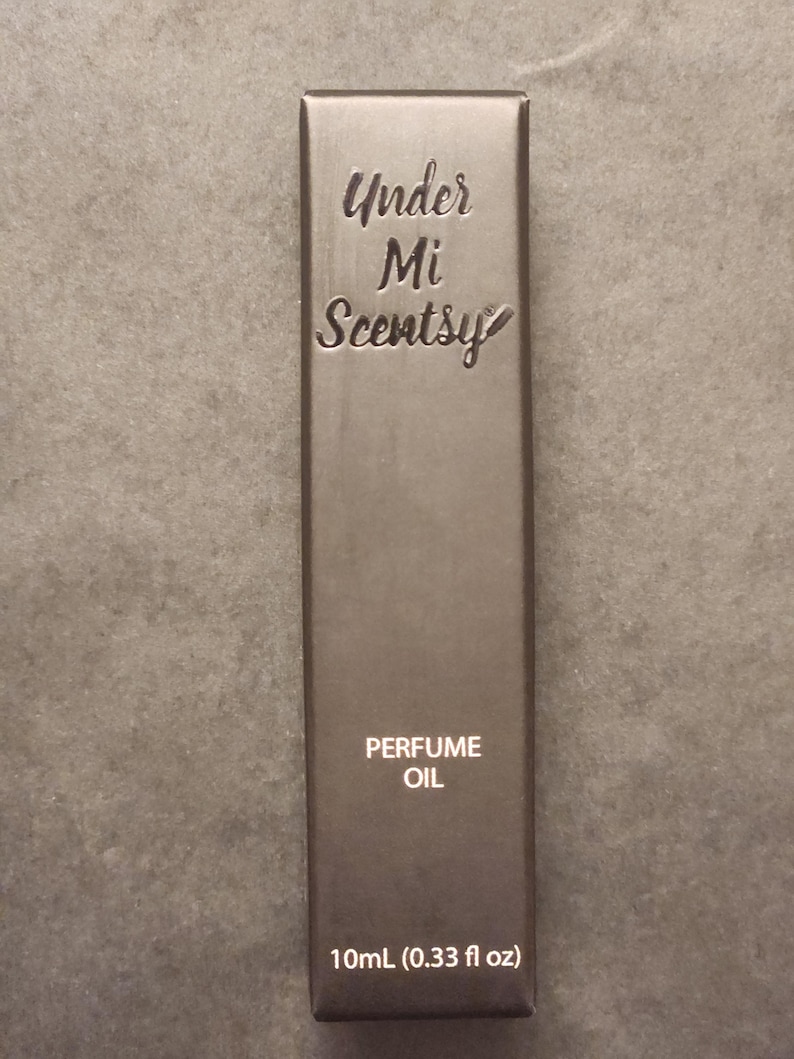 Puede incluir: Una caja rectangular de color marr&oacute;n oscuro para aceite de perfume. El frente presenta el texto en relieve Under Mi Scentsy en una fuente cursiva, seguido del texto impreso PERFUME OIL y la medida de volumen 10mL (0.33 fl oz) en la parte inferior.