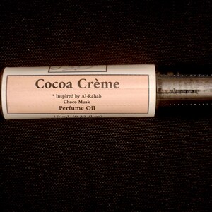 Puede incluir: Un frasco cil&iacute;ndrico de aceite de perfume con una etiqueta de color melocot&oacute;n claro que muestra el texto Cocoa Cr&egrave;me, inspired by Al-Rehab Choco Musk, Perfume Oil, y 10 ml (0,33 fl oz). El frasco tiene una tapa met&aacute;lica oscura y cuerpo de cristal.