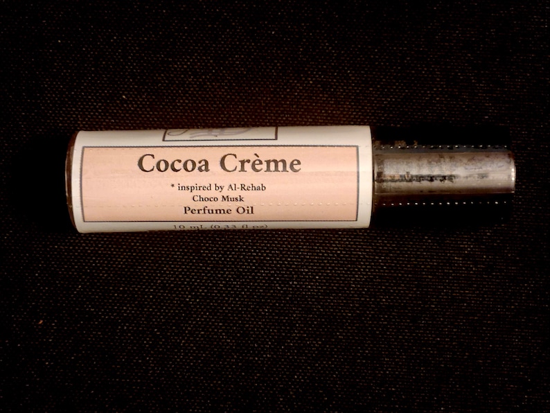 Puede incluir: Un frasco cil&iacute;ndrico de aceite de perfume con una etiqueta de color melocot&oacute;n claro que muestra el texto Cocoa Cr&egrave;me, inspired by Al-Rehab Choco Musk, Perfume Oil, y 10 ml (0,33 fl oz). El frasco tiene una tapa met&aacute;lica oscura y cuerpo de cristal.