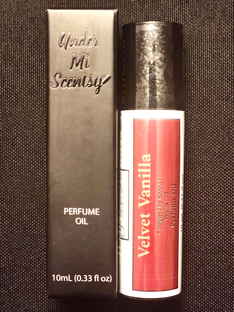 May include: A black rectangular perfume oil box labeled Under Mi Scentsy and 10mL (0.33 fl oz) next to a cylindrical perfume oil bottle with a black cap and a white label featuring the text Velvet Vanilla, inspired by KAYALI Vanilla | 28.
