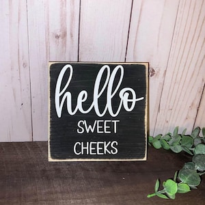 Peut inclure: Petit panneau carré en bois avec un fond noir et des lettres blanches. Le panneau indique "hello SWEET CHEEKS" dans un style manuscrit. Le panneau a une bordure marron clair et est posé sur un fond en bois.