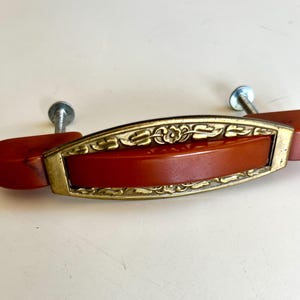 May include: A drawer pull featuring a central horizontal bar made of smooth, burnt-orange material encased in a decorative, antiqued gold-tone metal frame with floral and leaf engravings. Two silver-coloured metal mounting screws are attached.