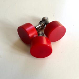 May include: Three circular red knobs with smooth, flat tops and metallic silver-coloured cylindrical stems. Each knob features a solid red finish and a short, sturdy metal base designed for attachment, presented as a small group of hardware components.