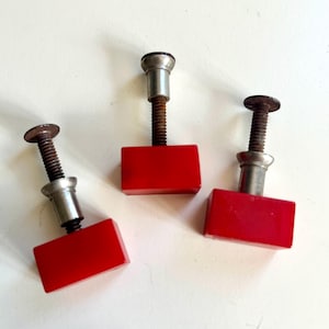 May include: Three rectangular cabinet knobs with a glossy red finish. Each knob features a metallic silver-toned cylindrical base and a threaded metal bolt showing signs of oxidation and rust, extending from the back of the red rectangular handle.