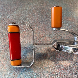 May include: A vintage hand-cranked kitchen tool featuring a chrome metal frame, a side-mounted rotating handle with an orange grip, and a main vertical handle composed of a central red cylinder flanked by two smaller orange cylindrical accents.
