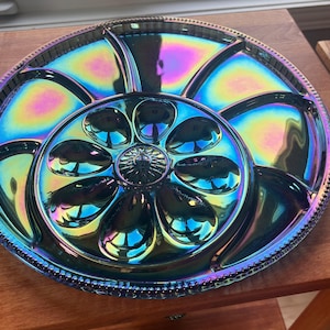 May include: A round iridescent serving platter with a metallic finish that shifts between blue, purple, and green hues. The tray features a central circular section with eight small, oval-shaped indentations surrounding a decorative centre point.