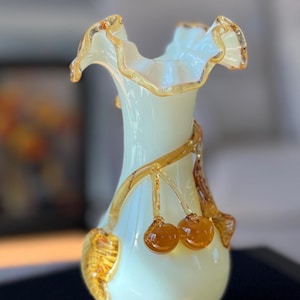 May include: A decorative glass vase with a frilled rim and a light green body. The vase is decorated with amber-coloured glass vines and two amber-coloured glass cherries. The vase has a round base and a slender neck.