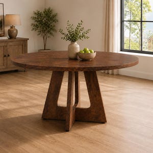 May include: A large, round wooden dining table with a textured, brown surface. The table is supported by a sturdy, geometric base. A vase with greenery and a bowl of green apples are on the table. The table is in a well-lit room.