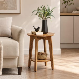 May include: A round wooden side table with four tapered legs and a lower shelf. The tabletop holds a black vase with green eucalyptus branches, a small black bird figurine, and a book. A small woven basket sits on the lower shelf.