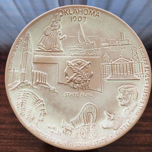 May include: A round, tan ceramic decorative plate featuring embossed illustrations of Oklahoma state symbols, including the state flag, a pioneer woman, a sailboat, a church, a covered wagon, a Native American figure, and Will Rogers with cattle.