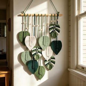 May include: A macrame wall hanging featuring multiple leaf-shaped fiber art pieces in various shades of green and cream. The leaves are suspended from a horizontal wooden dowel by knotted cords, with small wooden beads interspersed along the hanging strings.