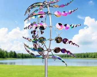May include: A multi-colored metal wind spinner with a reflective, iridescent finish. The garden art features circular and leaf-shaped elements arranged on a central pole. The background includes a green lawn, lake, and blue sky.