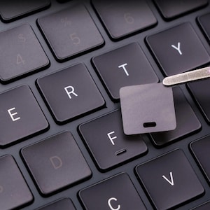 May include: A black computer keyboard with white letters including E, R, T, Y, D, F, C, and V. A pair of silver metal tweezers holds a small, square, blank black keycap replacement piece directly above the F key on the keyboard surface.