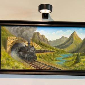 May include: A framed landscape painting depicts a steam train emerging from a tunnel, traveling through a mountain valley. The artwork features a black train, green hills, a blue lake, and a blue sky. The frame is black.