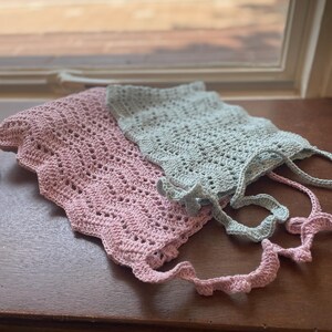 May include: Two crocheted items with a wavy, scalloped edge design. One item is a soft pastel pink color, and the other is a light mint green color. Both pieces feature an openwork stitch pattern and include long, thin crocheted ties or straps.